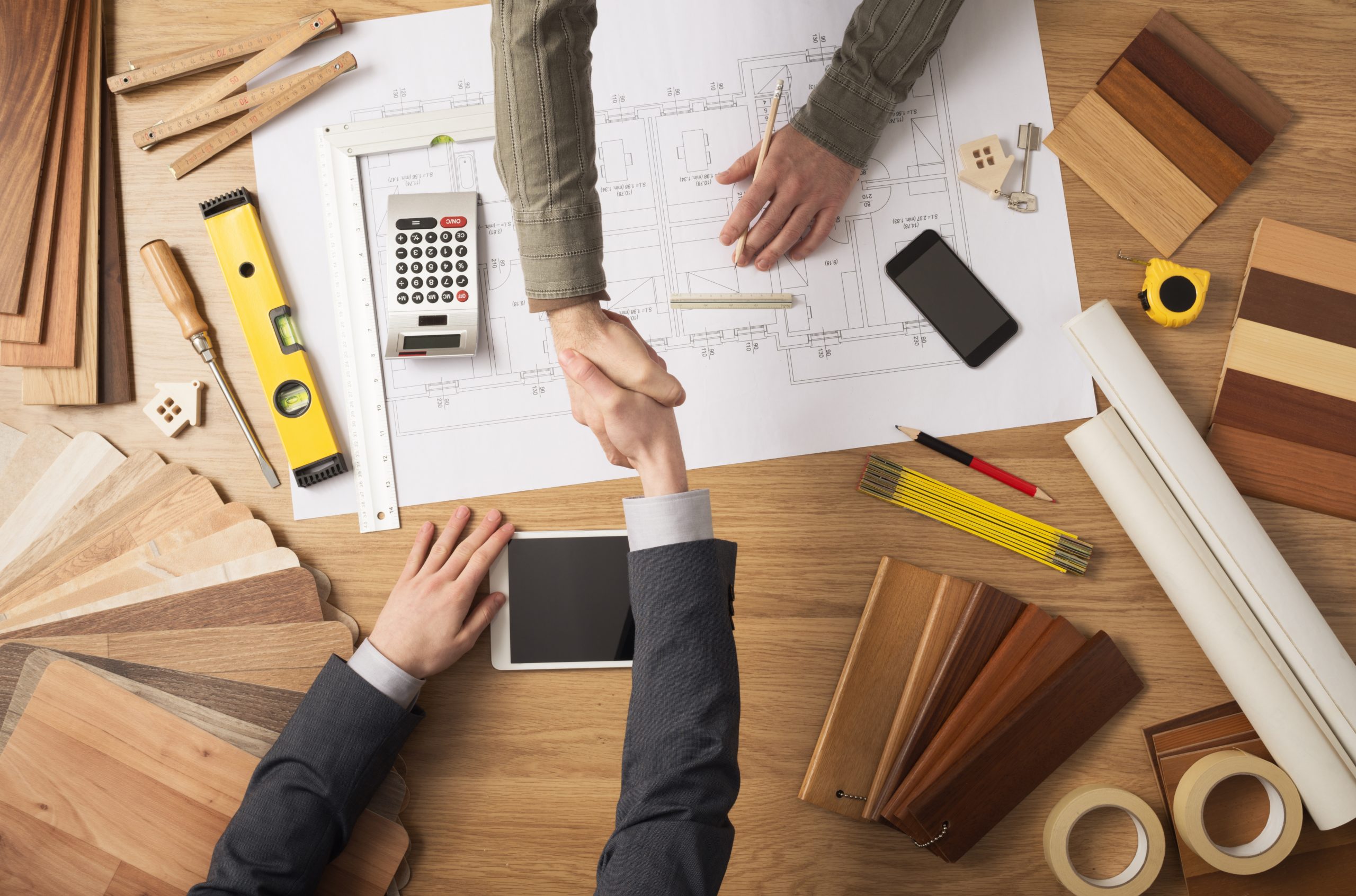 Architect and customer businessman shaking hands top view, desktop with building project, tools and wood swatches on background